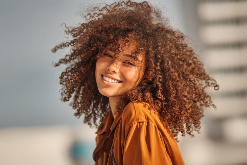 portrait of a beautiful young woman with curly hair smiling in the sunshine, showcasing natural beauty and carefree spirit against a blurred urban background.