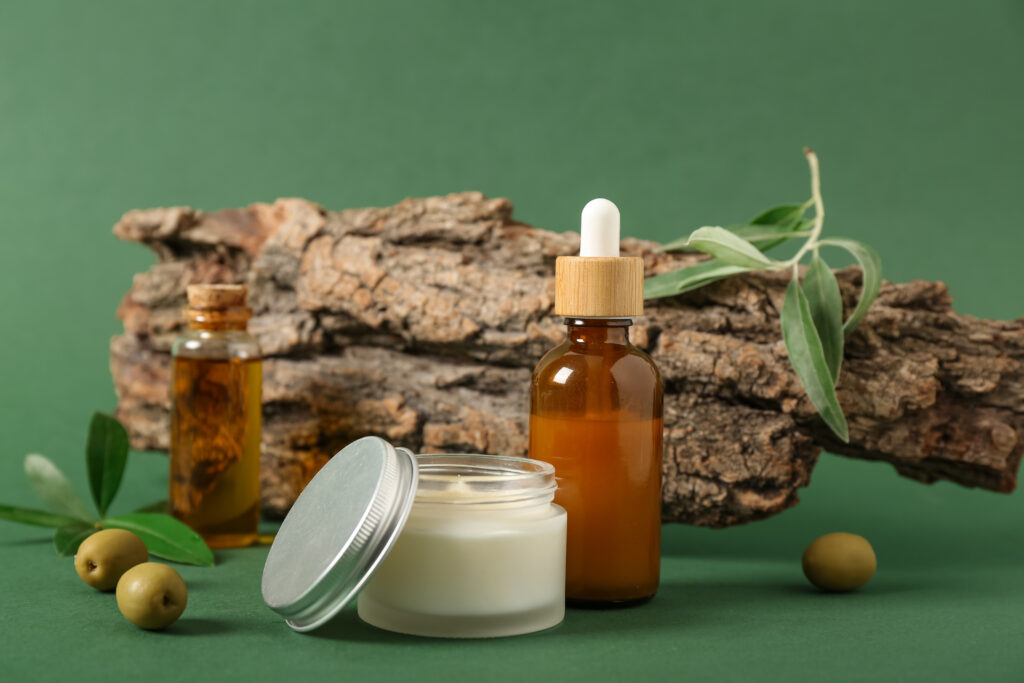 composition with tree bark, bottles of essential olive oil and jar of cosmetic product on green background, closeup