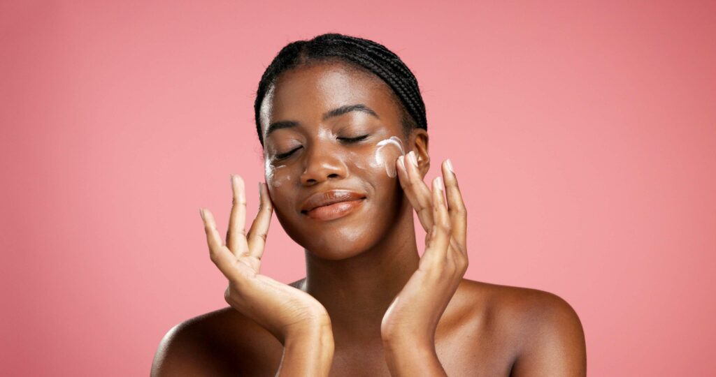 black woman, lotion and skincare with face cream in studio for moisturizer on a pink background. female person, model and applying cosmetics for skin protection, anti aging or sunscreen on space