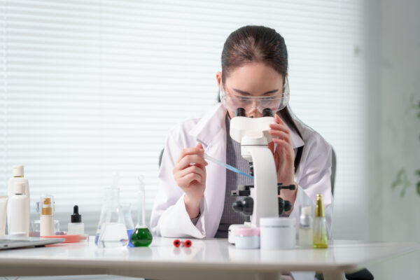 female scientist in lab coat and safety goggles using microscope at lab bench with glassware, droppers and cosmetic bottles, bright clean laboratory setting.