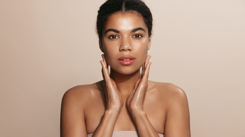 close up shot of dark skinned woman with curly hair, appiles face cream on cheek, poses nude against brown background, cares of beauty, uses cosmetic product. natural beauty concept